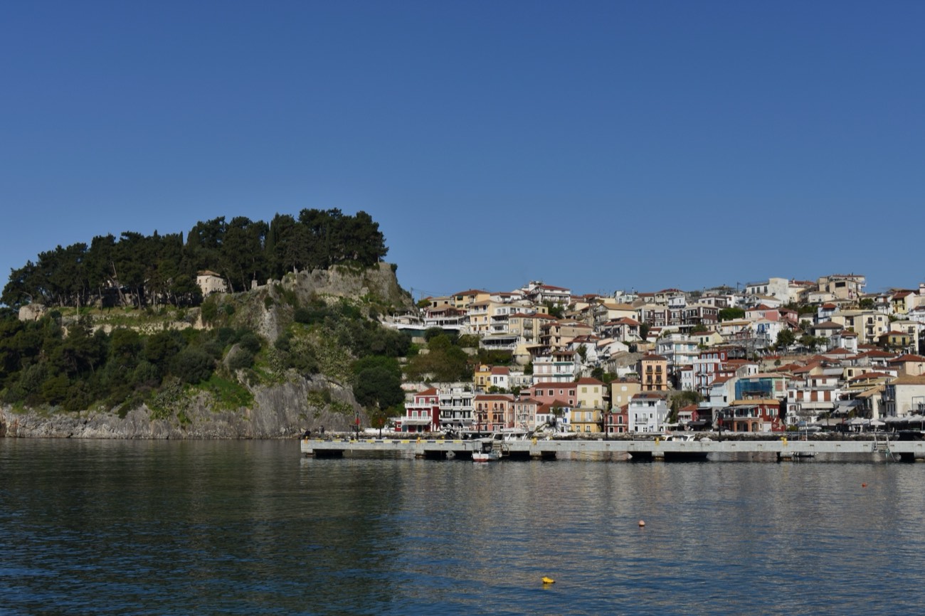 A view of Parga, Greece from the harbour, with the wooded, walled hill of Parga Castle on the left and the old-town houses climbing the hillside.