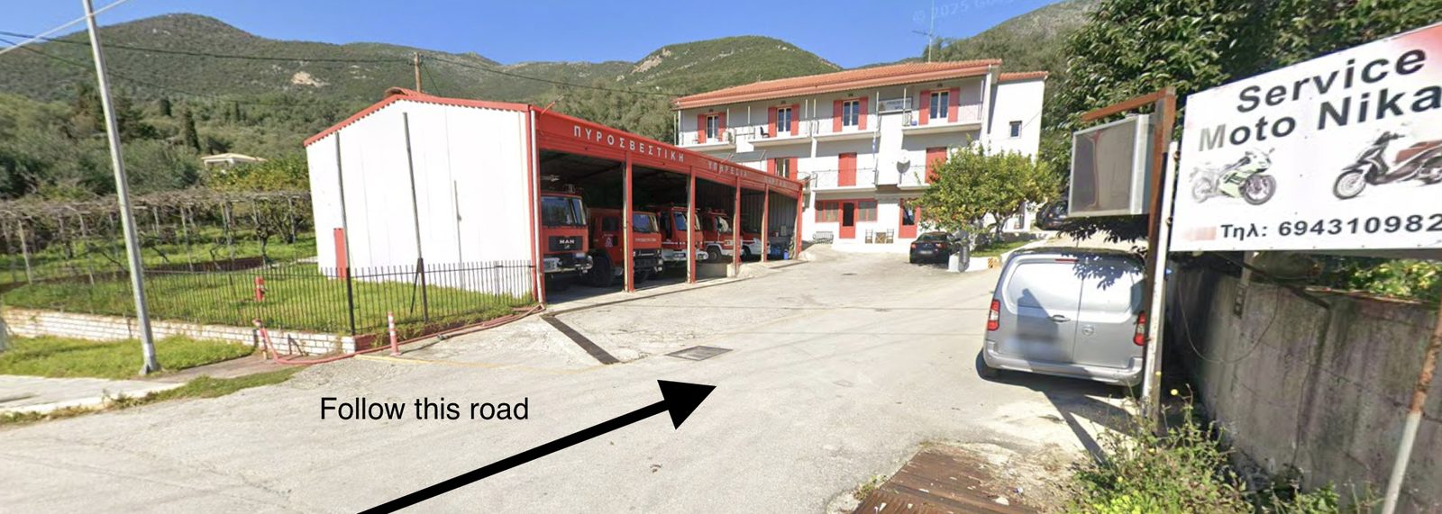 Parga fire station, a white building with red shutters and open bays holding fire engines, with an arrow along the road that passes in front of it